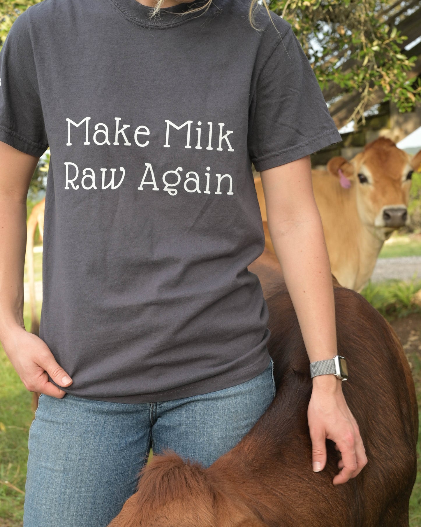 Person wearing a shirt with 'Make Milk Raw Again' text, standing next to a cow outdoors.