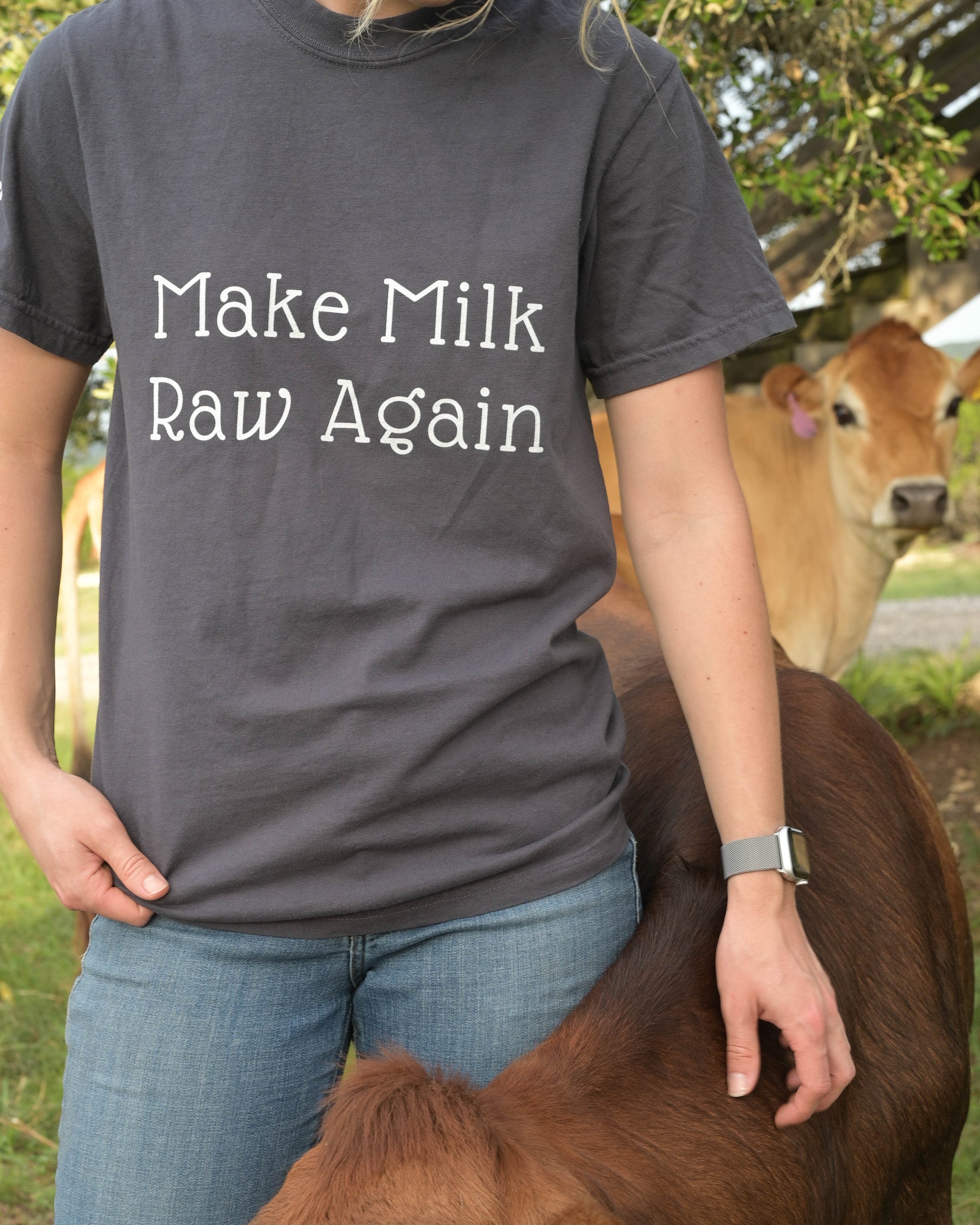 Person wearing a shirt with 'Make Milk Raw Again' text, standing next to a cow outdoors.
